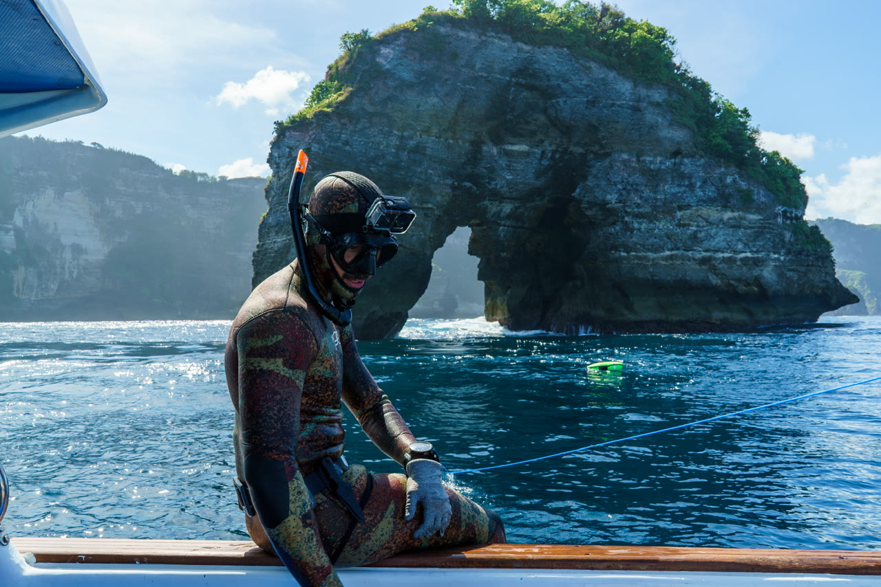 A spearfisher sits on the boat with a coastal rock arch behind him.