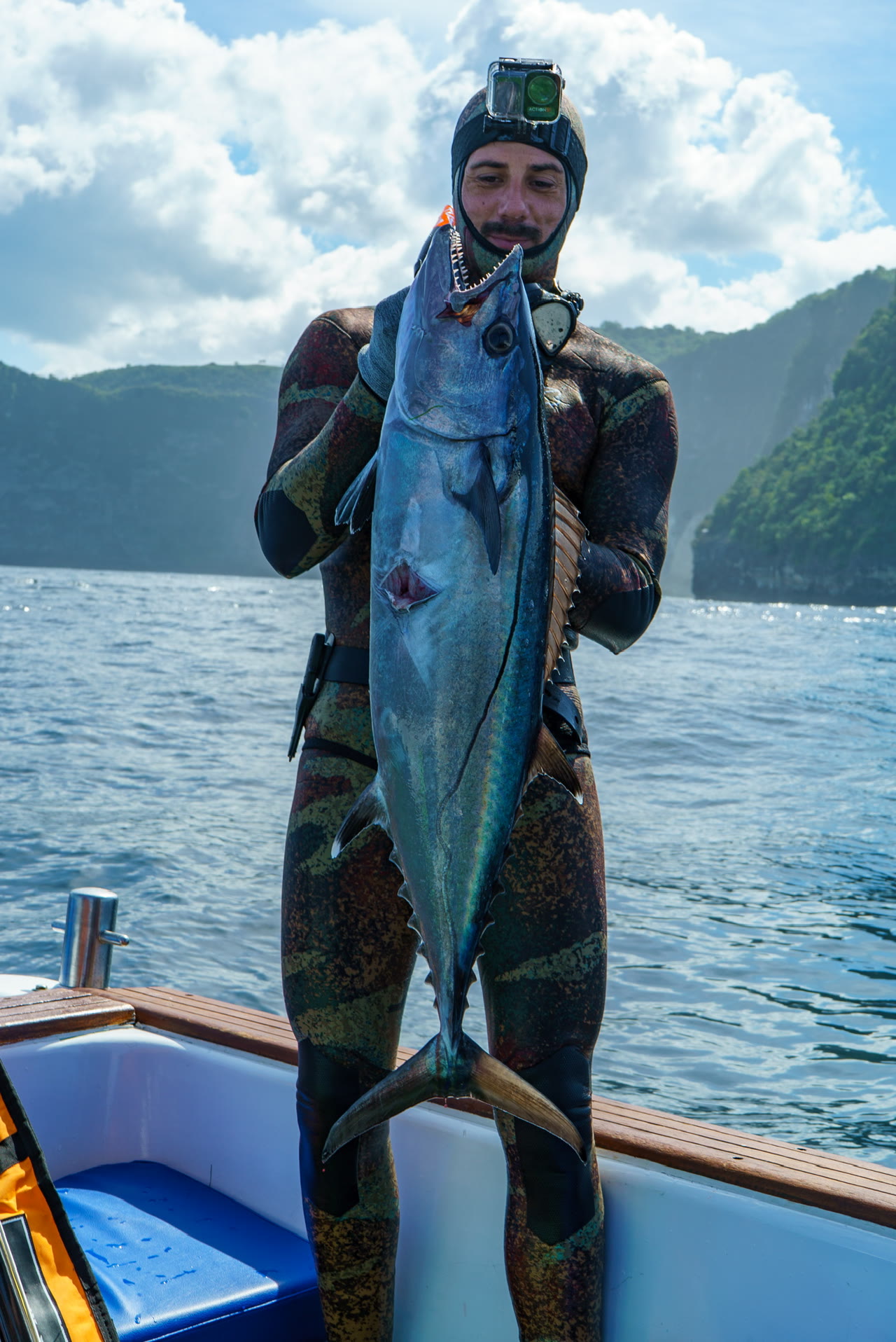 A smiling spearfisher holds a large silver catch with blue water and cliffs behind him.