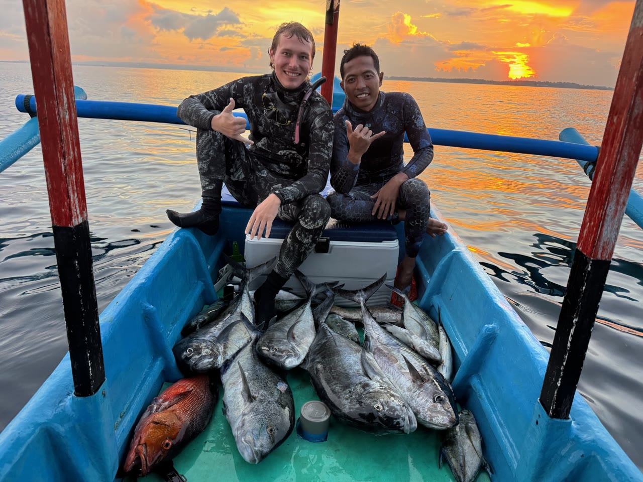 Two smiling spearfishers sit on a blue boat behind their catches at sunset.