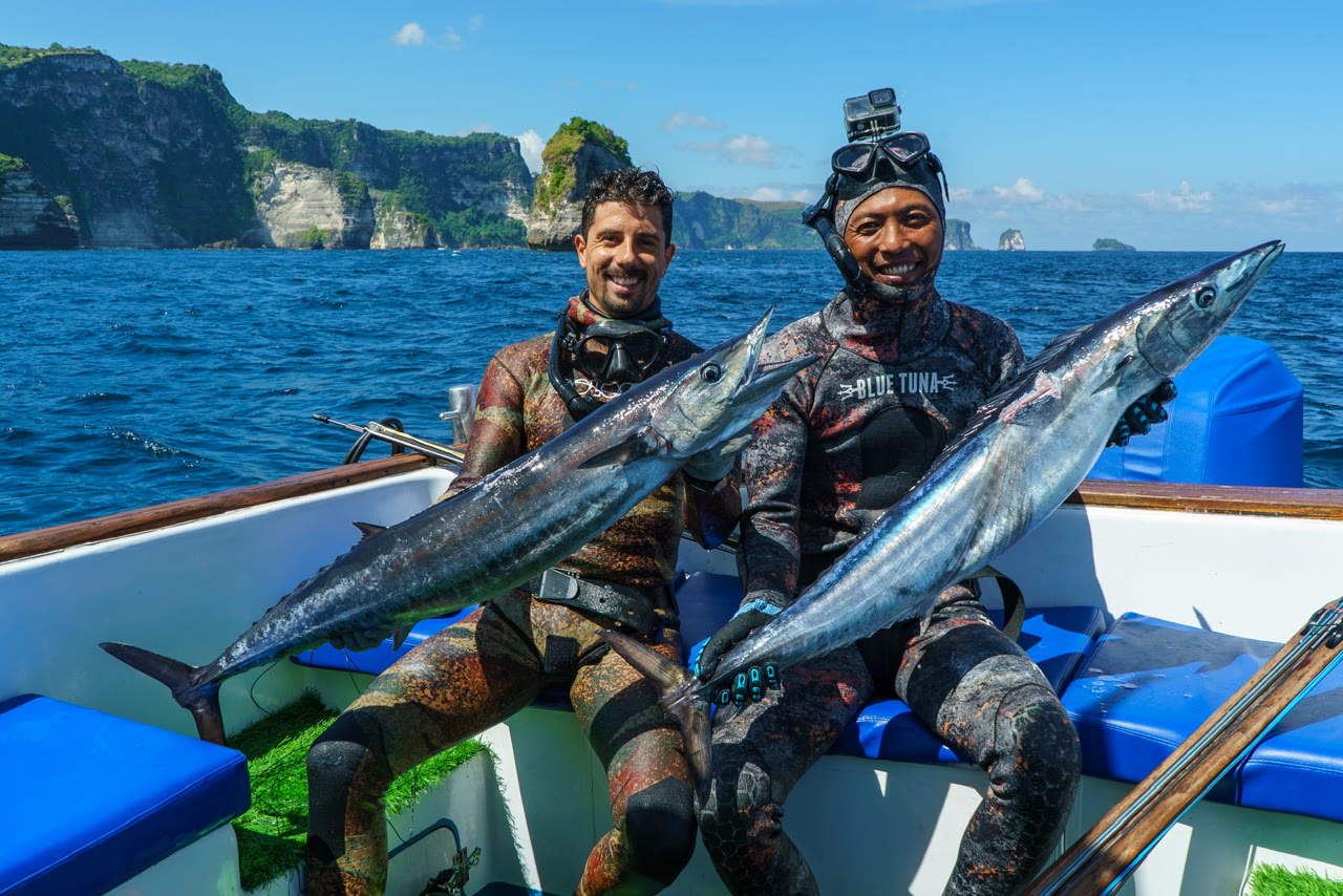 Two spearfishers sit aboard a blue boat holding their catches against a coastal backdrop.